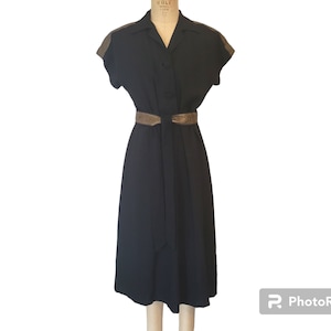 Może przedstawiać: Czarna sukienka z krótkimi rękawami, kołnierzykiem i pasującym paskiem. Sukienka posiada ozdobne akcenty na ramionach i zapinana jest na guziki z przodu. Pasek jest w metalicznym złotym kolorze.