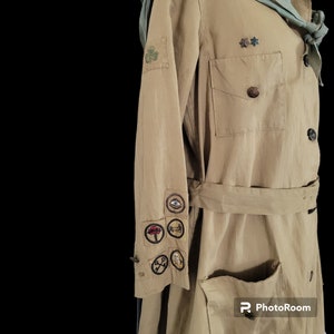 May include: A tan-colored utility jacket with a green sash and several embroidered patches on the sleeve. The patches feature various symbols, including an eye, a tree, a key, and a compass.