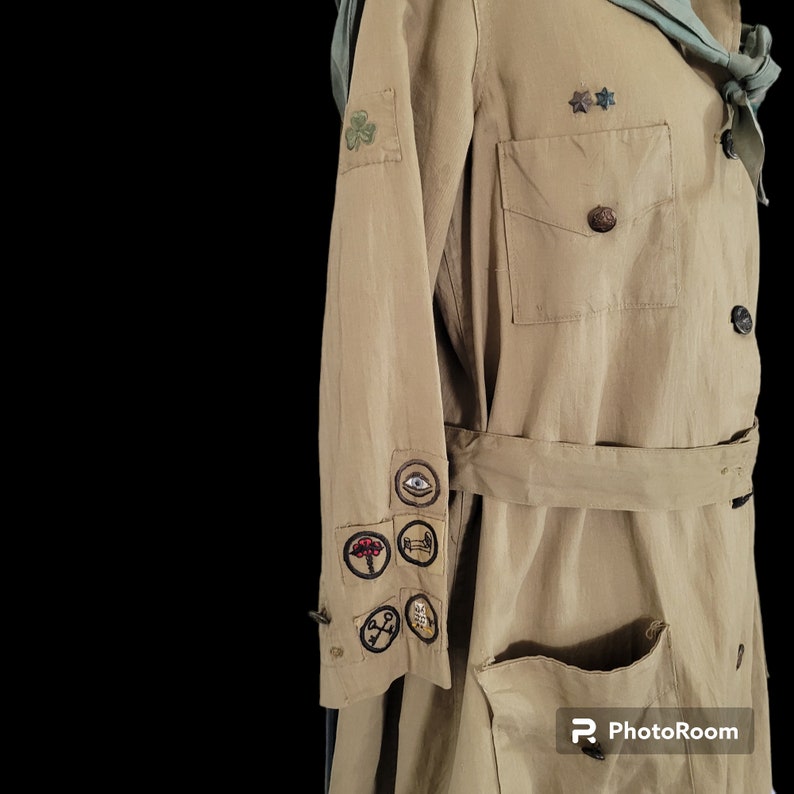 May include: A tan-colored utility jacket with a green sash and several embroidered patches on the sleeve. The patches feature various symbols, including an eye, a tree, a key, and a compass.