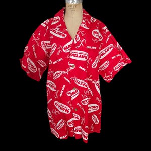 Puede incluir: Una camisa roja de manga corta con un estampado blanco en toda la superficie de la palabra "MAYTAG" y el lema "The Dependable Automatic".