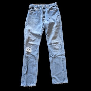 May include: Light blue denim jeans featuring a high-waisted fit, a button-fly closure, and a distressed rip on the right knee. The pants have a straight-leg silhouette with classic five-pocket styling, copper-toned rivets, and frayed hems.