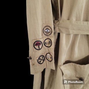 May include: A beige utility jacket with a variety of embroidered patches on the sleeve. The patches feature symbols like an eye, a tree, a bed, a key, and an owl.