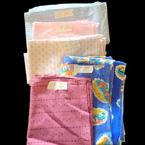 May include: A stack of six fabric scraps in various colors and patterns, including pink, blue, and white. Each scrap has a handwritten label indicating the amount of fabric in yards. The labels read "1 1/2 yds", "2 yds", "2 1/2 yds", "1/2 yds", "1/3 yds", and "6/7 yds".