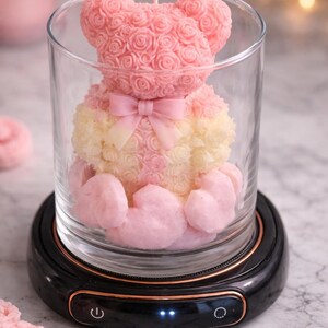 May include: A pink and yellow teddy bear candle inside a clear glass jar. The bear is made of rose-shaped wax and has a pink bow. The jar sits on a black base with copper-colored trim and control buttons.