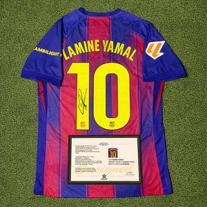 Puede incluir: Una camiseta de fútbol azul y roja con el nombre "LAMINE YAMAL" y el número "10" en amarillo. La camiseta tiene una firma y la palabra "AMBILIGHT" en la manga. Se muestra un certificado enmarcado debajo.