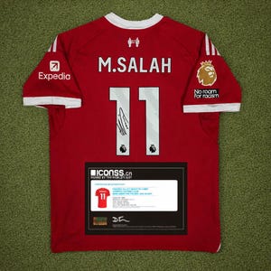 May include: A red Liverpool football jersey with the name "M. SALAH" and the number "11" on the back. The jersey has white accents and logos for Expedia and the Premier League. Includes a certificate of authenticity.