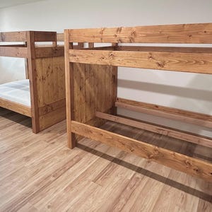 May include: Two wooden bunk beds with a natural finish. The beds have a simple design with a ladder and guardrails. The beds are made of solid wood and have a sturdy construction. The room has light-colored wood flooring and white walls.