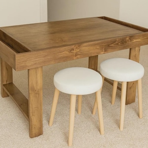 May include: A wooden table with a recessed top and two matching stools. The table is approximately 48 inches long and 30 inches wide. The stools have white padded seats and light-colored wooden legs.