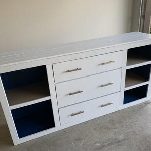 Puede incluir: Una cómoda de madera blanca con tres cajones y estanterías abiertas. La cómoda tiene tiradores dorados y un interior azul. La parte superior está hecha de tablones de madera.