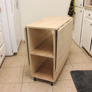 Puede incluir: Un carrito de cocina de madera clara sin terminar, con una tapa abatible y dos estantes interiores. El carrito tiene ruedas. Las dimensiones son aproximadamente 76 cm de largo, 51 cm de ancho y 76 cm de alto.
