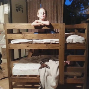 May include: A wooden bunk bed with a natural finish. The bed has two levels, each with a mattress and bedding. A child is sitting on the top bunk. The room has a kitchen and a window with curtains.