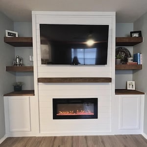 May include: A bespoke entertainment unit with a white shiplap design, incorporating a large flat-screen TV, a fireplace with an orange glow, and floating wooden shelves. The unit includes two white cabinets and is set against a light blue wall.