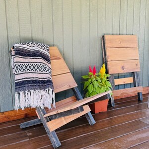 May include: A wooden Adirondack chair with a black and white patterned blanket draped over it. A small folding table and a potted plant with red and yellow flowers are nearby. The chair has a rustic, handmade appearance.