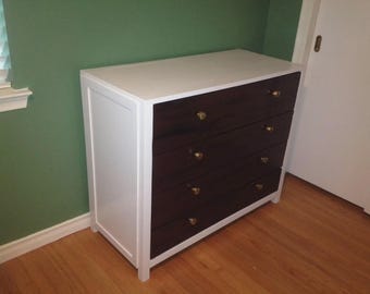 May include: A four-drawer dresser with a white frame and a dark brown drawer front. The top is white, and the drawers have gold-colored knobs. The dresser is placed against a green wall and a wooden floor.