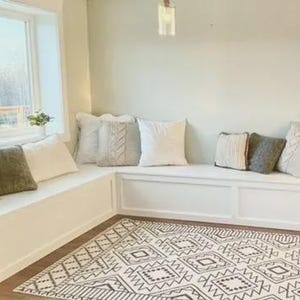 Può includere: Una panca bianca integrata con diversi cuscini decorativi in tonalità neutre. La panca è posizionata di fronte a una finestra e un tappeto fantasia è sul pavimento. I cuscini sono nelle tonalità del beige, grigio e bianco.