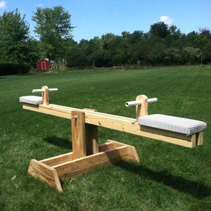 May include: A wooden seesaw with two seats, each with a striped cushion. The seesaw is set on a grassy lawn under a blue sky with a few clouds. The seesaw is made of natural wood and has a simple design.