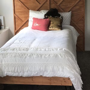 May include: A bed with a wooden headboard and frame. The headboard has a geometric design. The bed is made with white bedding, including a ruffled duvet cover and pillows. A pink pillow with the word "love" is visible.