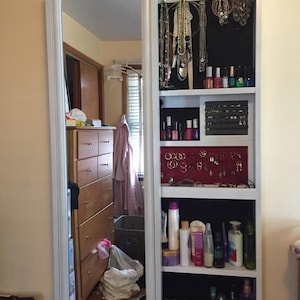 Può includere: Uno specchio a figura intera con cornice bianca con un armadietto nascosto per riporre gioielli e cosmetici. L'armadietto è aperto, rivelando ripiani pieni di collane, orecchini, smalto per unghie e vari prodotti di bellezza. Lo specchio è montato su una parete beige.