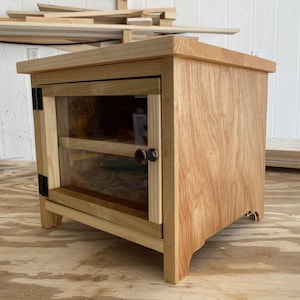 Może przedstawiać: Mała, drewniana szafka z przeszklonymi drzwiami. Szafka wykonana jest z jasnego drewna i posiada czarne zawiasy oraz ciemnobrązowe gałki. Szafka ma prostokątny kształt i stoi na krótkich nóżkach.