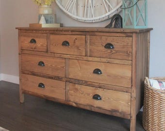 May include: A wooden dresser with seven drawers and black hardware. The dresser is made of light brown wood and has a rustic, farmhouse style. A large clock and decorative items are visible in the background.
