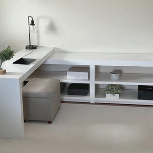 May include: A modern white desk with an attached shelving unit. The desk has a black lamp, a tablet, and a small plant. A gray ottoman is placed near the desk. The shelving unit displays decorative items and storage boxes.