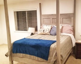 May include: A four-poster bed with a gray-washed wooden frame. The bed is made with a patterned comforter, a blue blanket, and several pillows. A window with blinds is visible in the background.