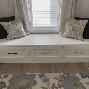 Puede incluir: Un banco de ventana blanco incorporado con tres cajones y manijas negras. Dos cojines decorativos con estampados florales y dos cojines verde oscuro se colocan en el asiento. La ventana está enmarcada por cortinas blancas y una alfombra estampada.