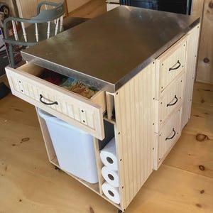 May include: A light wood kitchen island with a stainless steel countertop. The island has a drawer, a bin, and three drawers on the side. It also has a space for paper towels. The island is on wheels.