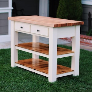 May include: A white kitchen island with a butcher block countertop and two drawers. The island has two lower shelves and black hardware. The wood shelves and countertop have a natural finish. The island is sitting on green grass.