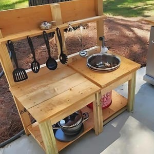 Puede incluir: Una cocina de juego de madera para exteriores con fregadero, estantes y utensilios colgantes. La cocina tiene un acabado de madera natural e incluye una olla de metal, un cubo rojo y varios utensilios de cocina negros. La cocina de juego está diseñada para niños.