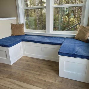 Puede incluir: Un asiento de ventana empotrado con gabinetes blancos y un cojín azul. Dos cojines decorativos marrones se colocan en el asiento. El asiento de la ventana está situado frente a una gran ventana con vistas a una zona boscosa.