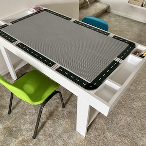 Puede incluir: Mesa de actividades de madera blanca con una superficie gris diseñada para bloques de construcción. La mesa tiene un diseño de carretera alrededor del borde y compartimentos de almacenamiento integrados. Una silla verde está colocada junto a la mesa.