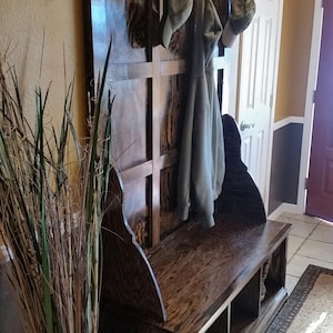 Könnte beinhalten: Dunkel gebeizte hölzerne Eingangsbank mit einer Rückwand mit Haken zum Aufhängen von Mänteln und Hüten. Die Bank hat einen eingebauten Stauraum und steht an einer Wand. Links steht eine Vase mit hohem Gras.