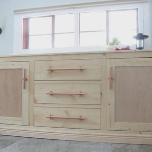 Op de afbeelding: Een lichtgekleurd houten dressoir met drie lades en twee kastdeuren. De lades en deuren hebben koperkleurige handgrepen. Het dressoir staat voor een raam en heeft een lichtgekleurde houten afwerking.