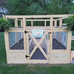 May include: A wooden greenhouse with a pitched roof, a door, and wire mesh windows. The door has an X-shaped brace and a sign that reads "Garden". Hanging baskets with ferns are on each side. A small wooden planter box is on the right.