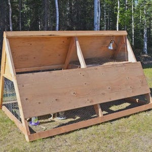 May include: A-frame chicken coop made of light-colored wood. The coop has a sloped roof and wire mesh sides and bottom. A small metal light fixture hangs inside. The coop is on a grassy lawn with trees in the background.