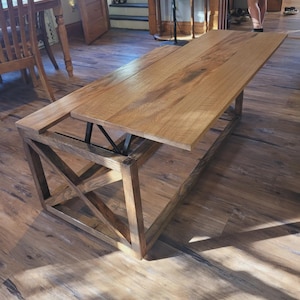 May include: A wooden coffee table with a lift-top design. The table has a natural wood finish, showing the grain. The base has an X-shaped design on the sides. The table is approximately 122 cm long, 61 cm wide, and 46 cm high.