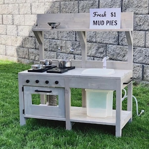 Pode incluir: Uma cozinha de brincar de madeira cinza para exterior com pia, fogão e forno. A placa acima da cozinha diz "Fresh $1 MUD PIES". A cozinha inclui panelas, frigideiras e uma torneira de água.