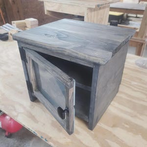 Może przedstawiać: Mała, szara, drewniana szafka z pojedynczymi uchylonymi drzwiami, odsłaniającymi puste wnętrze. Drzwi mają szklany panel i czarny uchwyt. Szafka stoi na jasnej drewnianej powierzchni.