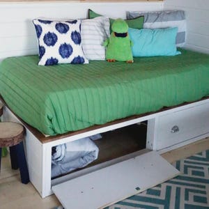 Puede incluir: Un sofá cama con una funda verde acolchada, cajones blancos y cojines decorativos. Dos pequeños taburetes de madera con tapas de madera natural y patas pintadas están frente a la cama. Un peluche verde está en la cama.