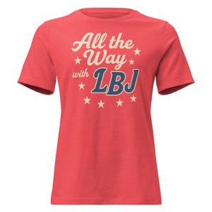 May include: A red t-shirt with the text "All the Way with LBJ" in a vintage style. The text is in cream and blue, with stars. The shirt has short sleeves and a crew neck.
