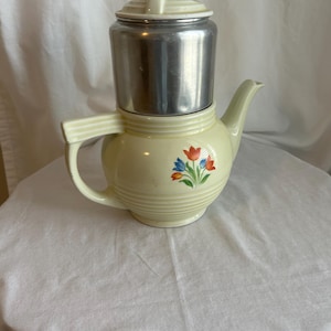 May include: A pale yellow ceramic teapot with a metallic silver top section and a matching pale yellow lid. The body features a floral decal with red, orange, and blue tulips. It has a side handle and a curved spout with horizontal ribbed detailing.