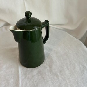 May include: A dark forest green ceramic pitcher with a matching lid and a side handle. The pitcher features a smooth, glossy finish, a curved spout for pouring, and a small knob on top of the lid. The interior of the spout is white.