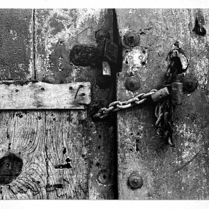 May include: A close-up of a weathered wooden door secured with a metal chain and padlock. The door is made of rough-hewn wood with a knot hole and is painted a faded gray. The chain is rusty and the padlock is old and worn.