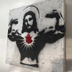 May include: A square canvas art piece featuring a black stencil of Jesus flexing his arms. A red heart with radiating lines is centered on the chest. The background is textured white.