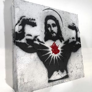May include: A canvas art piece featuring a black and white stencil of Jesus flexing his biceps. A red heart with rays emanates from his chest. The artwork has a textured, distressed appearance, with a white and gray background.