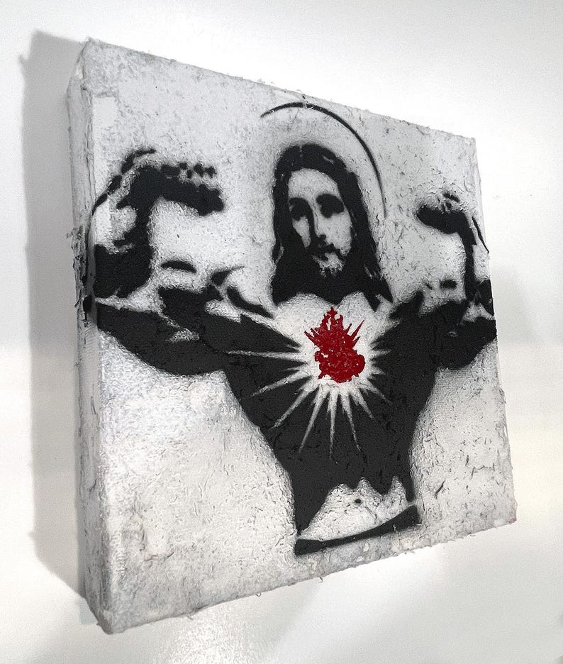 May include: A canvas art piece featuring a black and white stencil of Jesus flexing his biceps. A red heart with rays emanates from his chest. The artwork has a textured, distressed appearance, with a white and gray background.