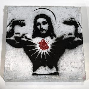 May include: A square, textured art piece featuring a black and white stencil of Jesus flexing his arms. A red, textured heart is in the center of his chest. A halo is above his head. The art piece has a white, textured background.