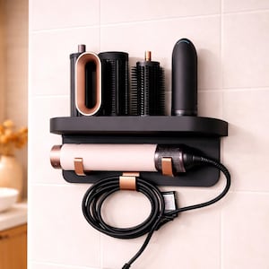 Peut inclure: Un organisateur mural noir pour outils de coiffure avec une étagère contenant divers accessoires. Un sèche-cheveux rose et cuivré est fixé en dessous. Le cordon d'alimentation et l'adaptateur noirs sont visibles. L'organisateur est fixé sur un mur carrelé.