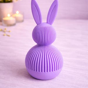 Könnte beinhalten: Ein lavendelfarbenes, hasenförmiges Deko-Objekt. Der Hase hat einen abgerundeten Körper und lange Ohren. Der Körper ist mit vertikalen Linien strukturiert. Der Hintergrund umfasst Kerzen und Blumen.